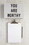`You are worthy` on a lightbox, clipboard with blank sheet of paper on a white wooden background, top view. Flat lay, overhead,