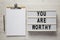 `You are worthy` on a lightbox, clipboard with blank sheet of paper on a white wooden surface, top view. Flat lay, overhead, fro