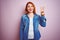 Youg beautiful redhead woman wearing denim shirt standing over isolated pink background showing and pointing up with fingers