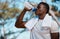Young active black male athlete holding a bottle and drinking water with a towel around his neck in a forest outside