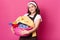 Young adorable smiling sweet female walking with pink basin in both hands, having much laundry, looks delighted and satisfied.