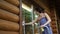 Young adult beautiful slim sporty caucasian woman in overalls stand on metal ladder and washing window wooden house