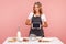 Young adult blonde woman in apron finds recipe on digital tablet, showing device with blank screen, cooking pastry, homemade