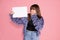 Young adult brown haired woman standing on pink background with whiteboard blank for sign on pink background in studio. Surpise,