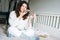 Young adult brunette woman forty years plus size body positive in white knitted sweater using mobile phone on the bed at home