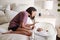 Young adult mother kneeling on her bed looking down at her three month old baby sleeping in his cot, side view