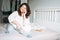 Young adult smiling brunette woman forty years plus size body positive in white knitted sweater using mobile phone on bed at home