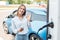 Young adult woman showing ok sign, standing near charging station for electric car