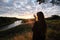 Young adult woman standing alone and staring at orange yellow sunset over river