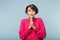 Young afraid woman with dark short hair in pink sweater keeping hands in praying gesture while amazedly looking aside