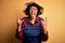 Young African American afro farmer woman with curly hair wearing apron and hat shouting with crazy expression doing rock symbol