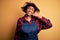 Young African American afro farmer woman with curly hair wearing apron and hat smiling doing phone gesture with hand and fingers