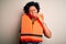 Young African American afro woman with curly hair wearing orange protection lifejacket touching mouth with hand with painful