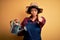 Young African American farmer woman with curly hair wearing apron and hat using water can pointing with finger to the camera and