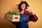Young African American farmer woman with curly hair wearing apron holding box with plants with surprise face pointing finger to