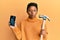 Young african american girl holding broken smartphone showing cracked screen and hammer making fish face with mouth and squinting