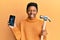 Young african american girl holding broken smartphone showing cracked screen and hammer sticking tongue out happy with funny