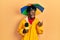 Young african american journalist man wearing yellow raincoat and umbrella cap pointing thumb up to the side smiling happy with