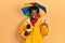 Young african american journalist man wearing yellow raincoat and umbrella cap surprised with an idea or question pointing finger