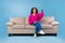 Young African American lady with remote control sitting on couch and watching TV on blue studio background, copy space