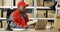 Young African American mailman in red uniform and cap sitting at desk in postal office store and working at laptop