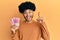 Young african american man with afro hair holding 100000 indonesian rupiah smiling with an idea or question pointing finger with