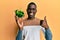 Young african american man holding bowl with small green peppers smiling happy and positive, thumb up doing excellent and approval