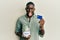 Young african american man holding credit card and jar with dollars sticking tongue out happy with funny expression