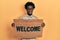 Young african american man holding welcome doormat in shock face, looking skeptical and sarcastic, surprised with open mouth