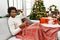 Young african american man sitting on the sofa drinking coffee by christmas tree strong person showing arm muscle, confident and