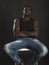 Young African-American man sitting on white chair on black isolated background.