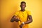Young african american man wearing casual t-shirt standing over  yellow background In hurry pointing to watch time,