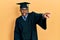 Young african american man wearing graduation cap and ceremony robe pointing with finger surprised ahead, open mouth amazed