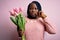 Young african american plus size woman with braids holding bouquet of pink tulips flower mouth and lips shut as zip with fingers