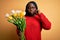 Young african american plus size woman with braids holding bouquet of yellow tulips flower mouth and lips shut as zip with fingers