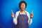 Young african american woman shop owner wearing business apron over blue background amazed and surprised looking up and pointing