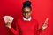 Young african american woman using smartphone holding norwegian krone banknotes clueless and confused expression