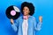 Young african american woman wearing headphones holding vinyl disc pointing thumb up to the side smiling happy with open mouth