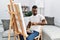 Young african man painting on canvas at home checking the time on wrist watch, relaxed and confident