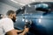 Young African man working at car detailing service, polishing blue car with polisher to eliminate contaminants from the