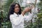 Young african woman biologist in white coat pouring clear liquid from glass flask into test tube, working in greenhouse