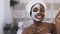 Young afro american woman in white towel on head applied facial clay mask on half face while holding grapefruit halves