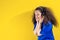 Young afro-haired girl listening to music with headphones on isolated yellow background