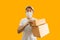 Young Asian delivery man wearing cap and white blank t-shirt holding parcel post box isolated on yellow background. express