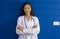 Young asian doctor in white gown and stethoscope stand with arm cross in front of blue wall