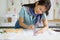 Young asian dressmaker in striped scarf use a pencil to draw cloth pattern on tracing paper. Tape measure, silk thread color