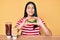 Young asian girl eating a tasty classic burger smiling looking to the side and staring away thinking