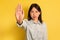 Young Asian lady gesturing STOP, showing palm at camera on yellow studio background