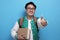 Young Asian male student wearing green baseball jacket holding books, smiling and shows thumbs up