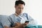 Young asian man with arm in sling lying on hospital bed looking at mobile phone. Broken arm patient